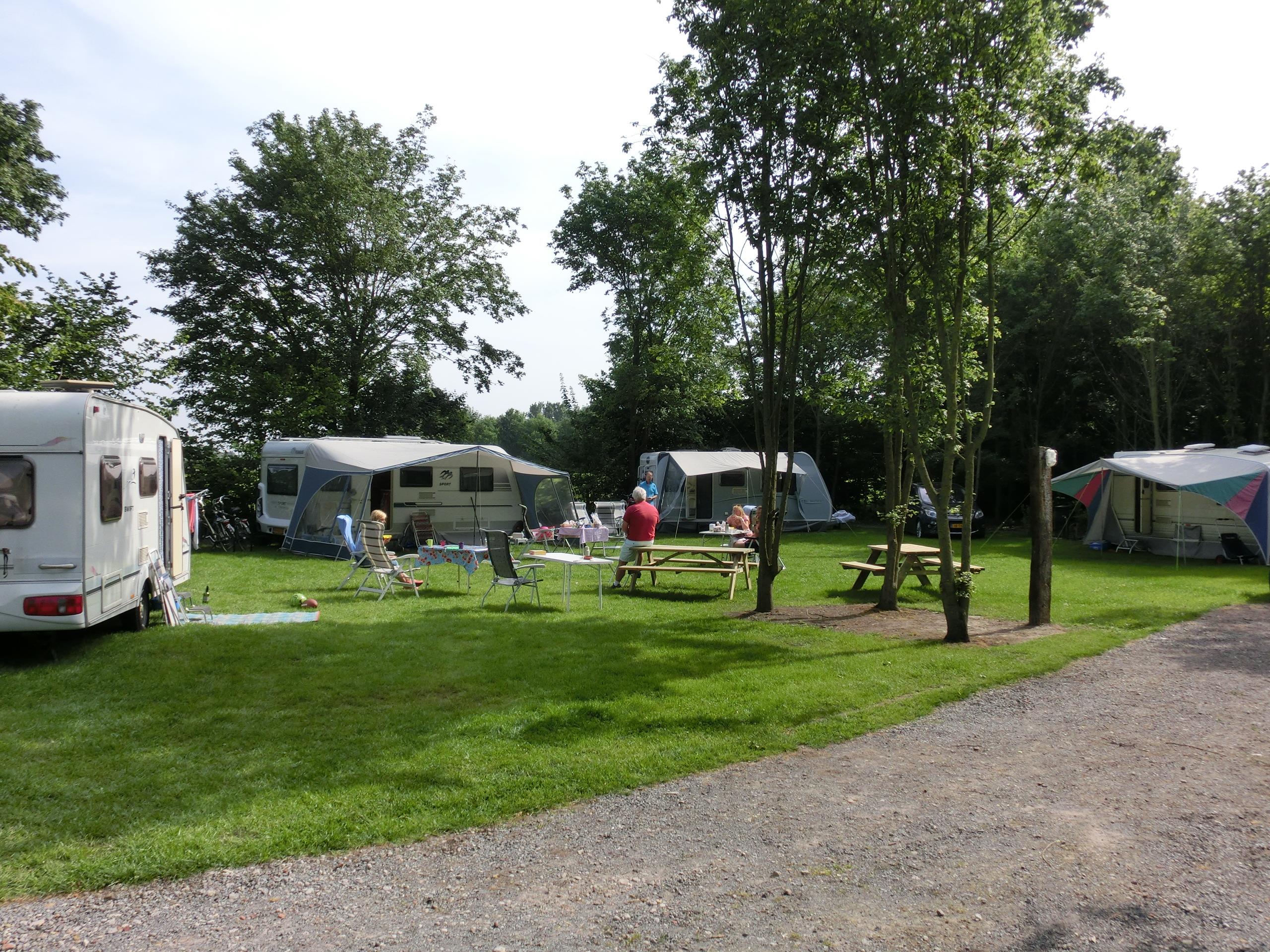 Campingplatz mit Wohnwagen und Zelten auf grünem Rasen, Sitzgarnituren und Bäumen entlang eines Kieswegs.
