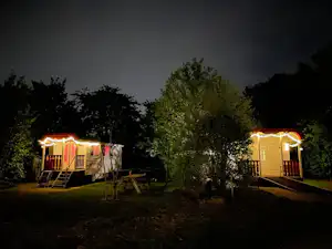 Zwei beleuchtete Mobilheime mit Lichterkette am Abend, umgeben von Bäumen und Rasen auf dem Campingplatz.