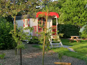 Blick auf ein Wohnmobil mit rotem Vordach und Veranda, Sitzbank und Tisch im Garten zwischen Bäumen und Kiesweg.
