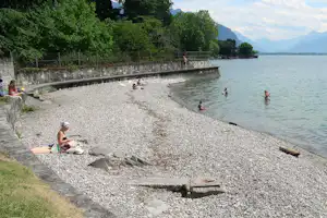 Kiesstrand am See mit Badebereich, Personen im Wasser und am Ufer, dahinter eine Steinmauer und Bäume.