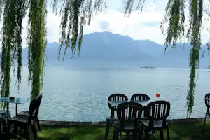 Campingplatz am See: Tisch und Stühle am Ufer, Weidenzweige im Vordergrund, Berge im Hintergrund.