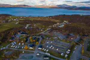 Luftaufnahme eines Campingplatzes am See mit vielen Wohnwagen und Häusern, Straßen und Bergen im Hintergrund.