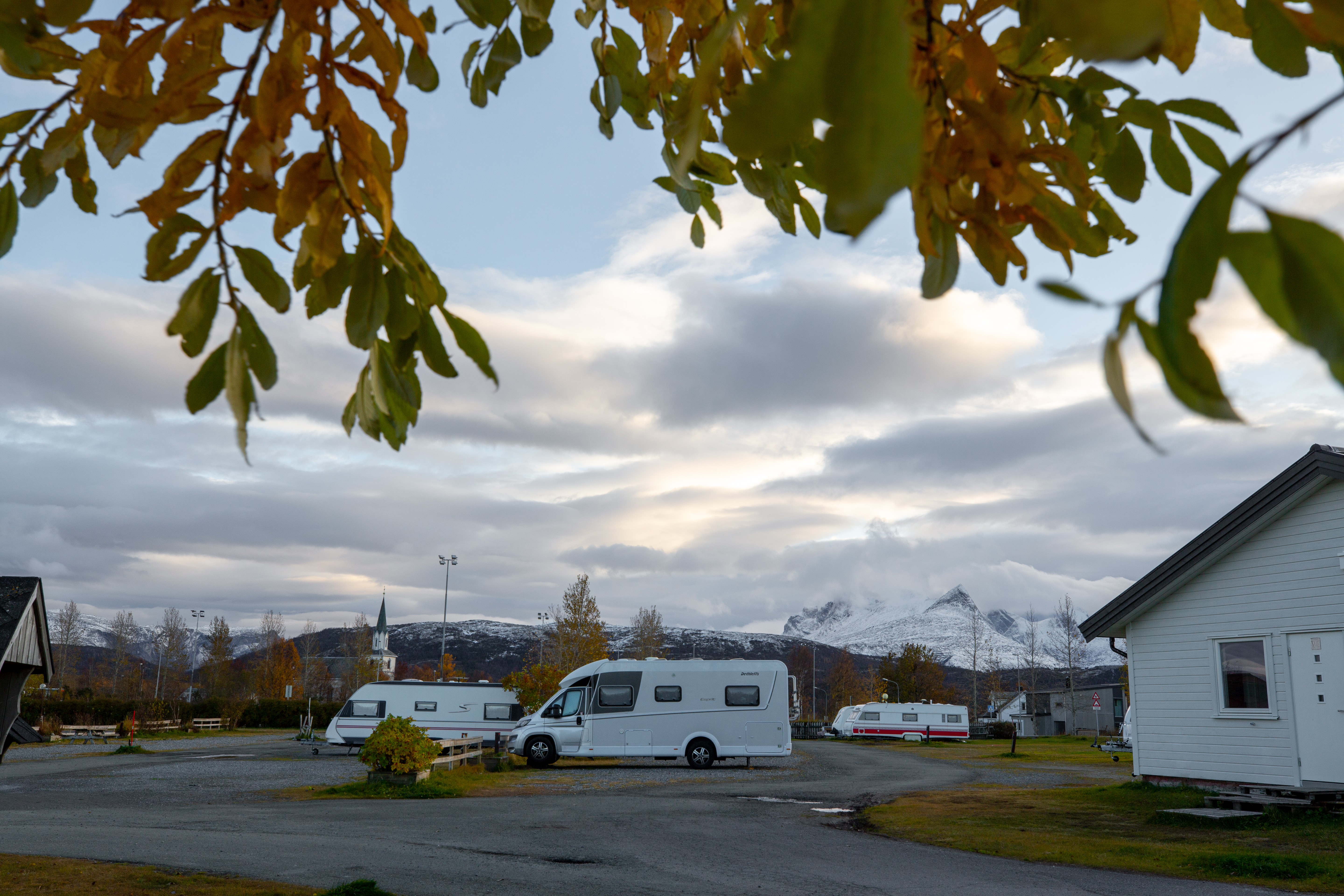 Wohnmobile und Wohnwagen auf einem Campingplatz, mit Gebäuden und Straßen im Hintergrund, unter Wolken und Bäumen.
