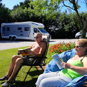 Zwei Personen sitzen auf Gartenstühlen im Campinggarten, im Hintergrund ein Wohnmobil auf dem Stellplatz und Bäume.
