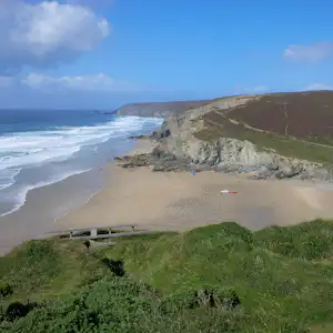 Weite Küstenansicht mit Sandstrand, Wellen und Kreidefelsen, im Vordergrund grünes Gras und ein kleiner Steg.