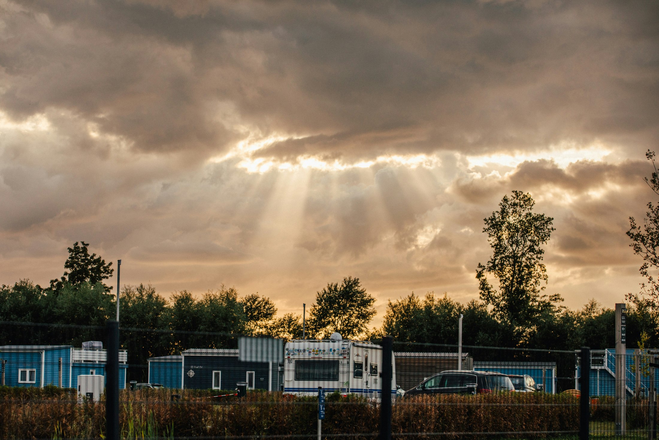 Blaue Container und Wohnwagen auf einem Campingplatz, im Hintergrund Bäume und ein bewölkter Himmel mit Sonnenstrahlen.