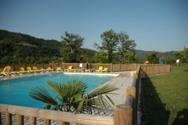 Poolbereich mit Liegestühlen, Steinmauer und Palmen im Garten eines Campingplatzes, dahinter Bäume und Hügel.