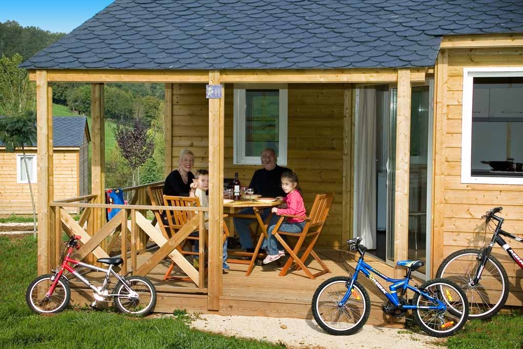 Holzhaus mit Terrasse, zwei Fahrräder davor und Sitzgruppe mit Tisch; im Hintergrund Bäume und weitere Gebäude.
