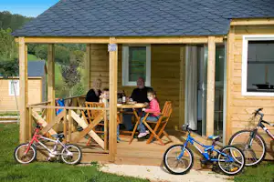 Holzhaus mit Terrasse, zwei Fahrräder davor und Sitzgruppe mit Tisch; im Hintergrund Bäume und weitere Gebäude.