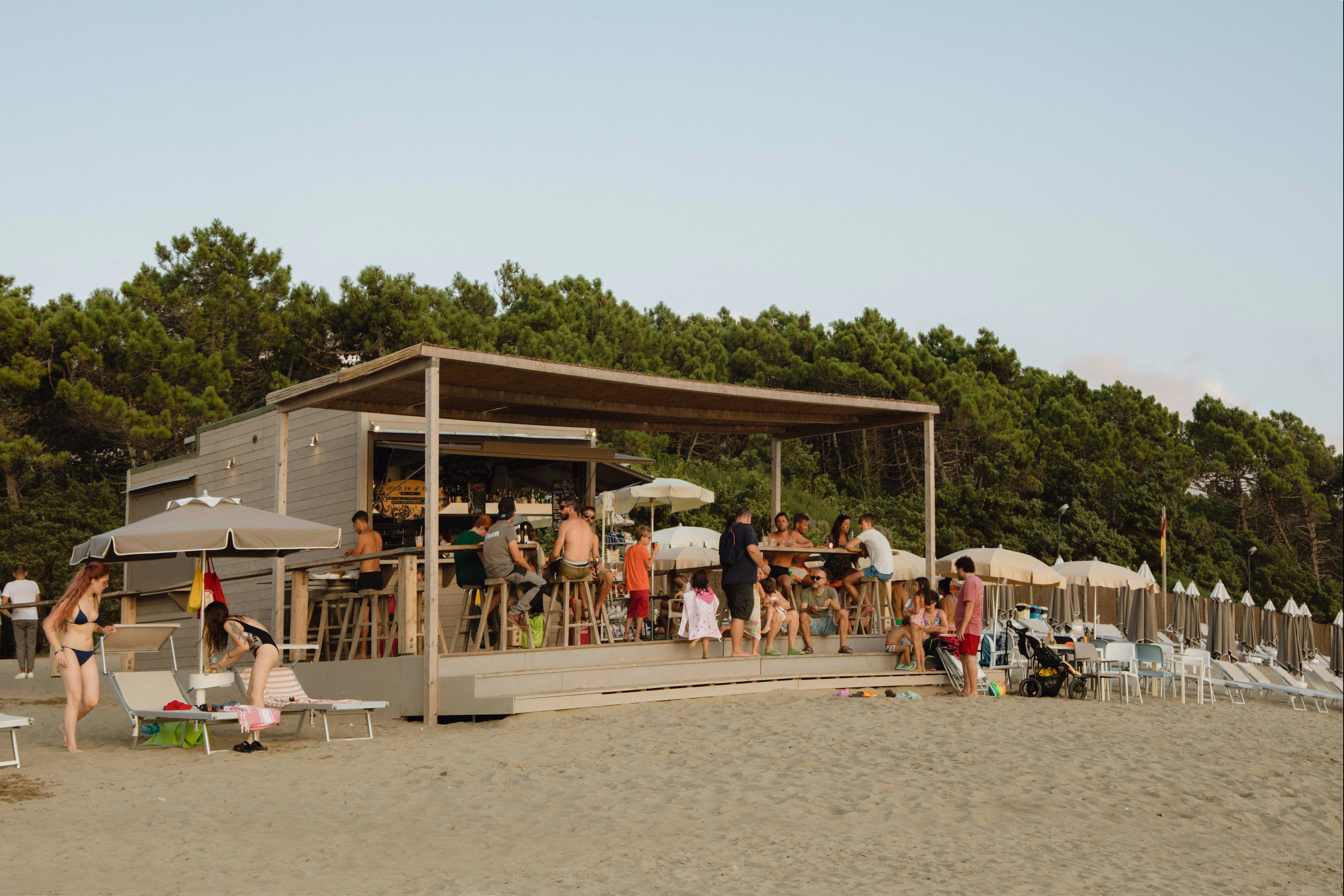 Holzgebäude mit Campingbar, viele Gäste an der Theke, Sonnenschirme und Stühle am Strand, im Hintergrund Kiefernwald.