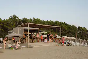 Holzgebäude mit Campingbar, viele Gäste an der Theke, Sonnenschirme und Stühle am Strand, im Hintergrund Kiefernwald.