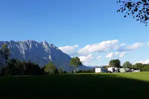 Grüne Wiese am Campingplatz mit Zelten und Wohnwagen, im Hintergrund Berge und Bäume unter klarem Himmel.