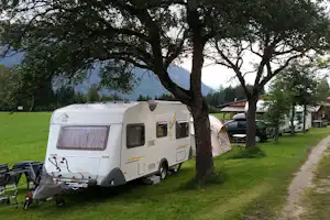 Wohnwagen auf einem Campingplatz-Stellplatz neben einem Grasweg, Bäume und ein See im Hintergrund.