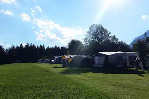 Campingwiese mit mehreren Zelten und Wohnwagen, Bäume am Rand und Berge im Hintergrund.