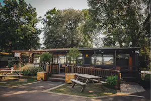 Holzhütten und eine Terrasse mit Sitzbank und Tisch auf einem Campingplatz, umgeben von Bäumen und Wegen.