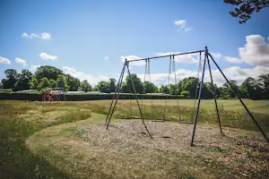 Schaukeln auf einem Spielplatz am Campingplatz, umgeben von Rasen, Bäumen und einem offenen Feld im Hintergrund.