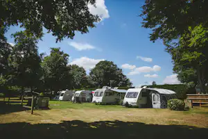 Campingplatz mit mehreren Wohnwagen und Zelten auf einer Rasenfläche, umgeben von Bäumen und Hecken.