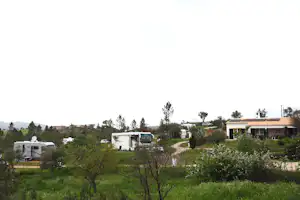 Wohnwagen und Campingbusse auf einem grünen Campingplatz, daneben ein Haus mit Terrasse, Bäume und Zufahrtsweg.