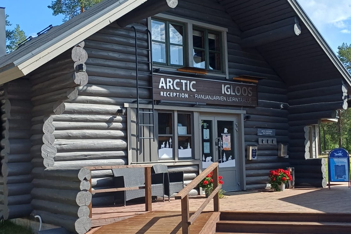 Blockhütte mit Eingang und Holztreppe, Schild „ARCTIC“ an der Rezeption, Terrasse mit Sitzbank und Blumen vor dem Gebäude.