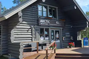 Blockhütte mit Eingang und Holztreppe, Schild „ARCTIC“ an der Rezeption, Terrasse mit Sitzbank und Blumen vor dem Gebäude.