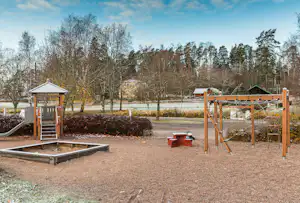 Spielplatz auf dem Campingplatz: Sandkasten, Holzschaukel und Rutsche, gepflasterter Weg und Bäume im Hintergrund.