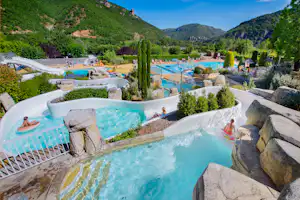 Wasserpark mit großen Pools, Wasserspielen und Liegestühlen, umgeben von Hecken und Bergen im Hintergrund.