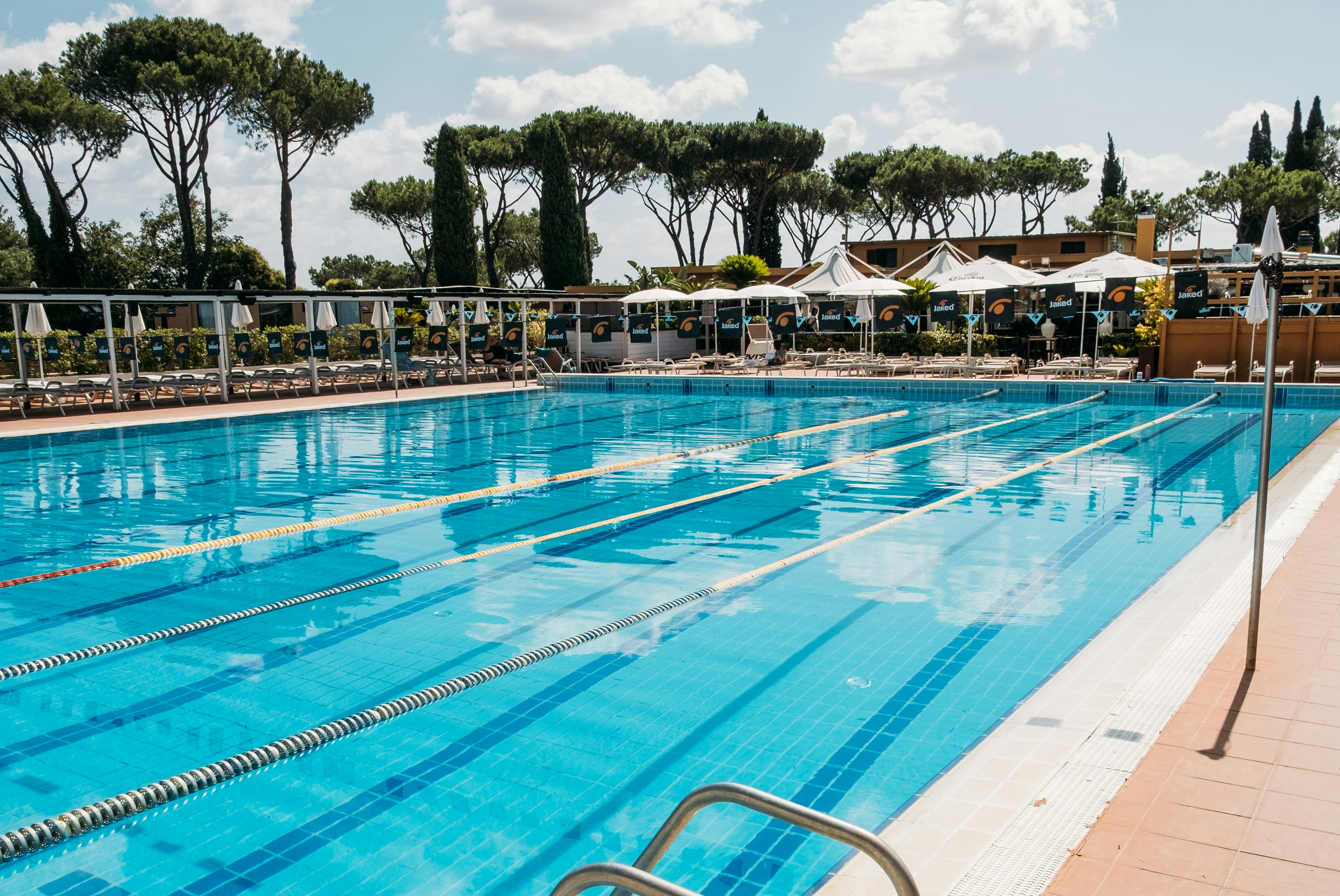 Schwimmbecken mit blauen Bahnen, Liegen und Sonnenschirmen am Campingplatz, im Hintergrund Bäume und Zäune.