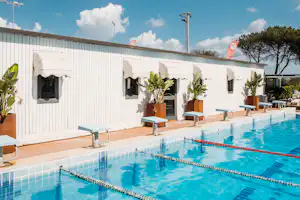 Pool mit blauem Wasser, Liegen und Terrasse vor weißen Bungalows mit Markisen und Fenstern.