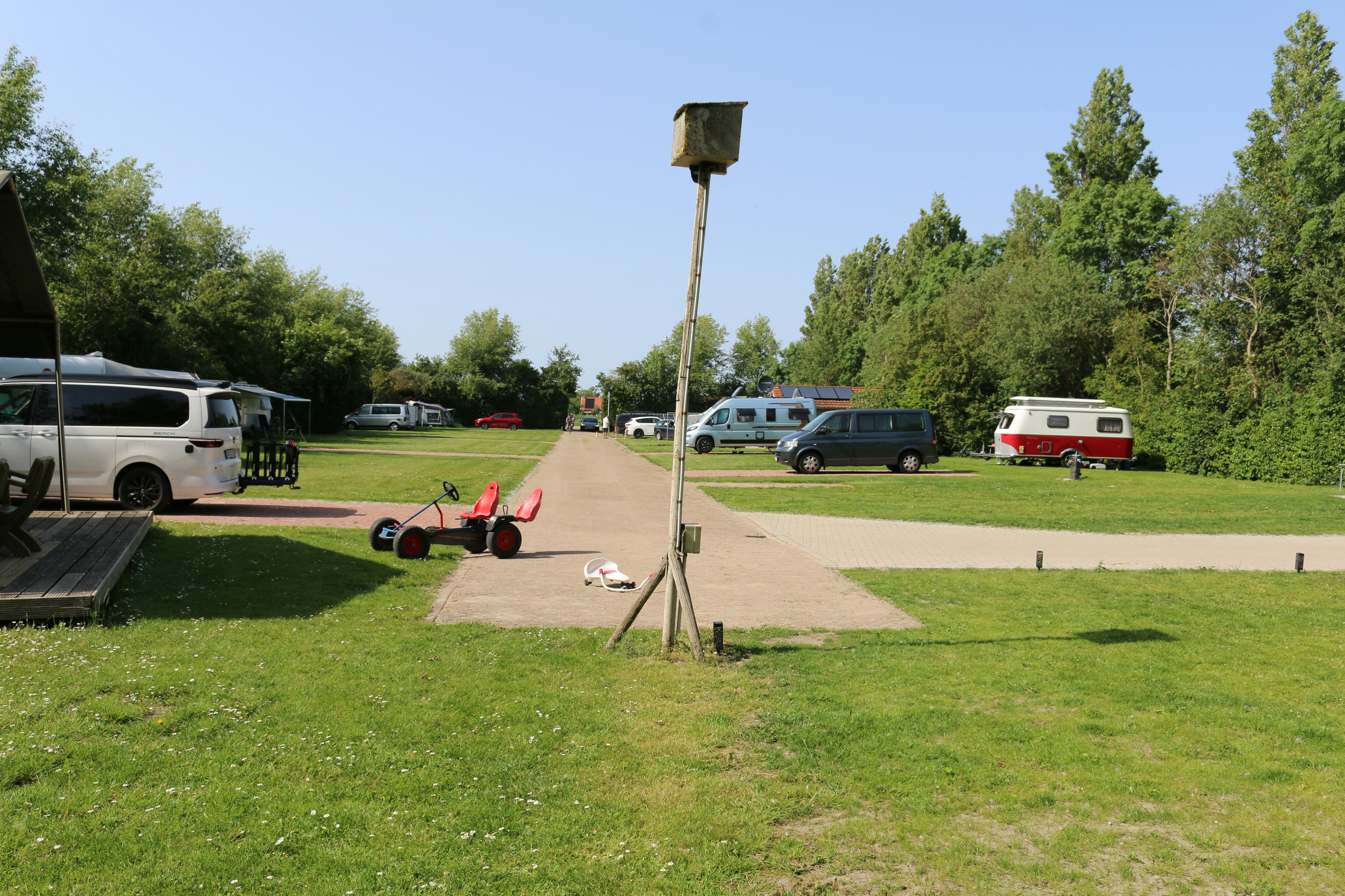 Stellplätze auf einem Campingplatz mit Rasen, Wohnmobilen und einem Weg; im Vordergrund ein Sonnenschirm und Campingstühle.