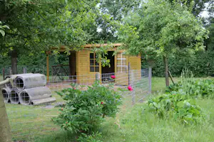 Holzunterstand mit Zaun und Sitzbank im Garten, daneben zwei gerollte Matten auf dem Rasen, umgeben von Bäumen.