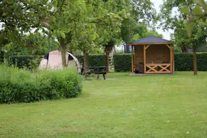 Stellplatz mit Wohnwagen und Holzunterstand, Tisch und Bank auf der Wiese, umgeben von Bäumen und Hecke.