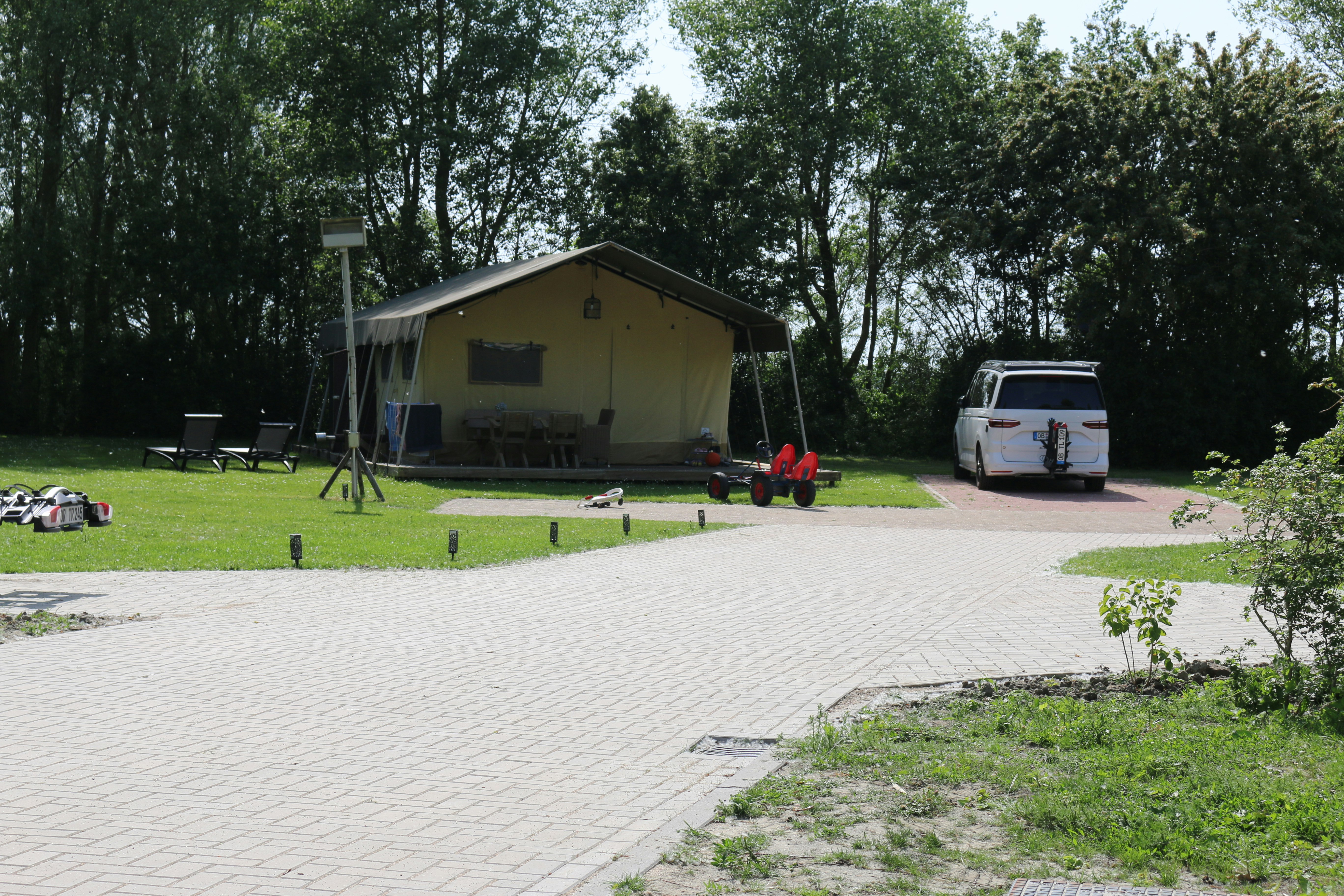 Gelbes Campinghaus mit Veranda im Grünen, daneben ein weißer Transporter und ein gepflasterter Weg durch den Campingplatz.