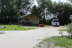 Gelbes Campinghaus mit Veranda im Grünen, daneben ein weißer Transporter und ein gepflasterter Weg durch den Campingplatz.
