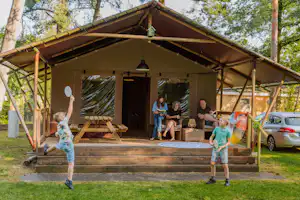 Familien-Campingzelt mit Holzterrasse, Sitzbank und Tisch, im Hintergrund Auto und Bäume im Campingplatzbereich.