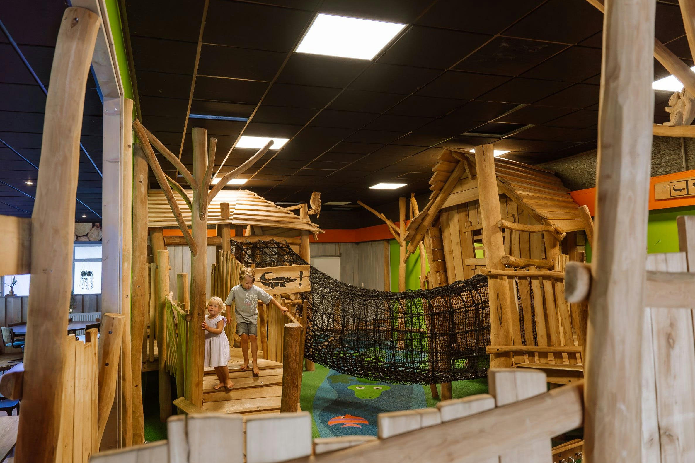 Holz-Spielbereich mit Klettergerüst, Netz und Rutschen im Innenbereich, mit buntem Boden und Kindern.