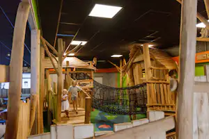 Holz-Spielbereich mit Klettergerüst, Netz und Rutschen im Innenbereich, mit buntem Boden und Kindern.