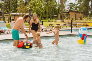 Familie im Schwimmbecken mit Spielzeugboot und Wasserspielzeug, im Hintergrund Liegen und Bungalows im Campingpark.