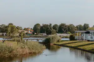 Wohnwagen und Mobilheime am Teich, Holzsteg und Ufergras mit Bäumen im Hintergrund.
