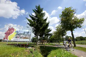 Campingweg mit Bäumen und Rasen, daneben ein großes Banner mit Text und Symbolen.