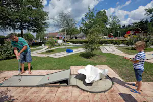 Minigolfanlage auf dem Campingplatz: zwei Kinder spielen an einer Bahn, im Hintergrund Bäume und kleine Gebäude.