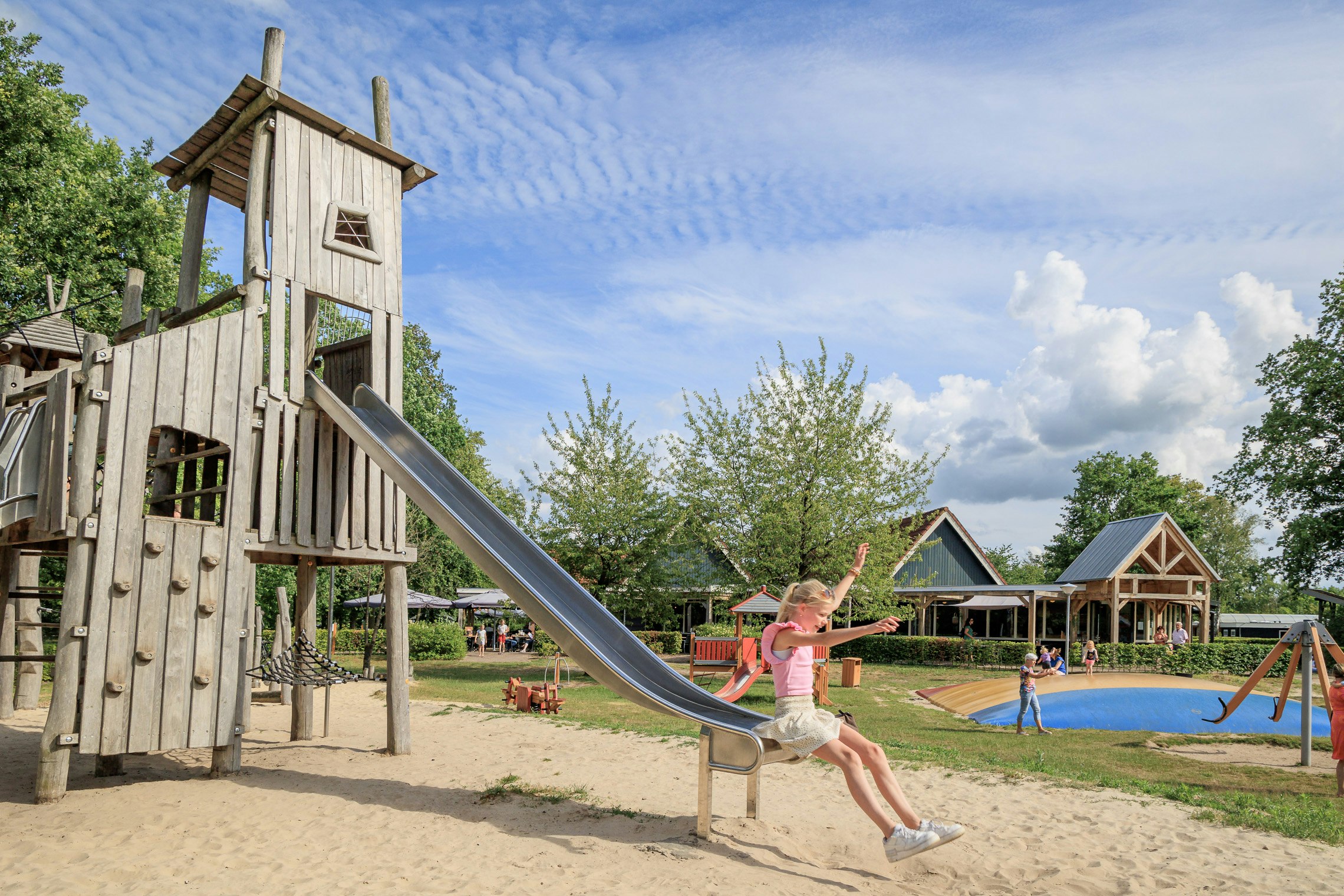 Holz-Spielturm mit Rutsche auf dem Campingplatz, im Hintergrund Spielbereich, Bäume und Gebäude unter blauem Himmel.