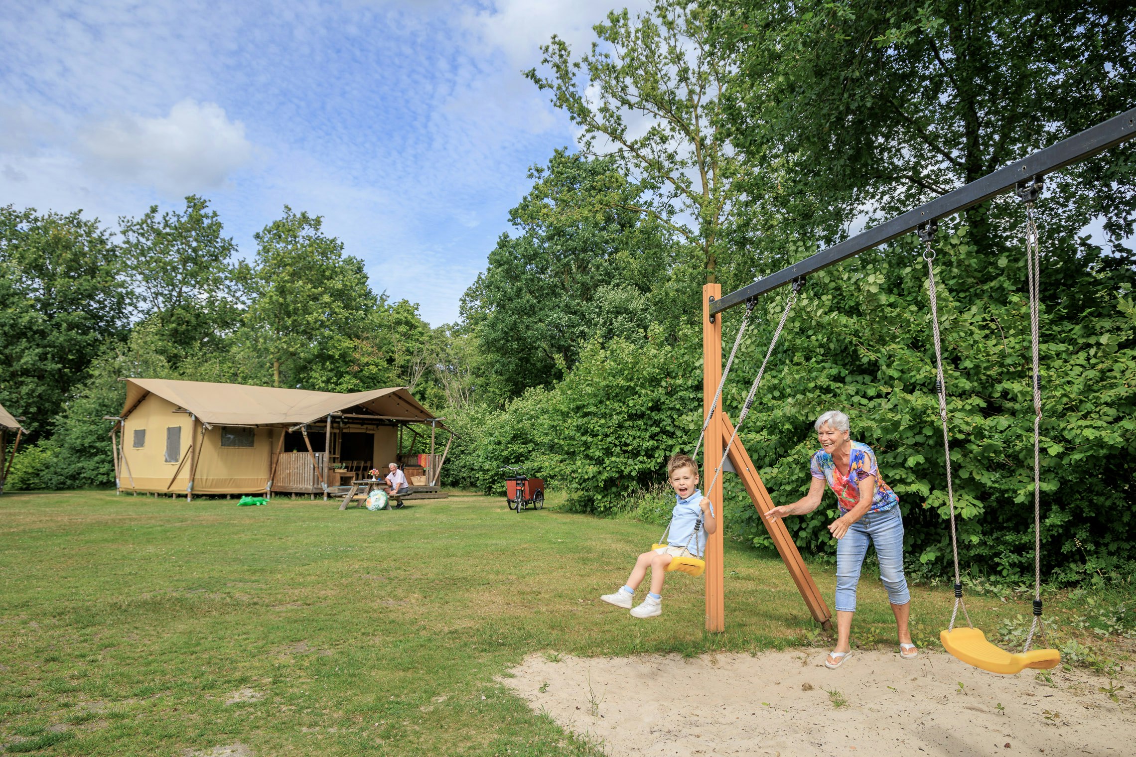 Familie auf einer Schaukel im Campinggarten, Zelt mit Terrasse im Hintergrund, viel Grün und Bäume.
