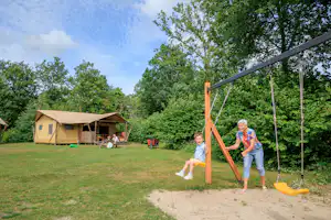 Familie auf einer Schaukel im Campinggarten, Zelt mit Terrasse im Hintergrund, viel Grün und Bäume.