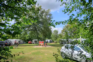 Campingplatz mit Zelten, Wohnmobil und Autos auf dem Stellplatz, umgeben von Bäumen und Rasenfläche.