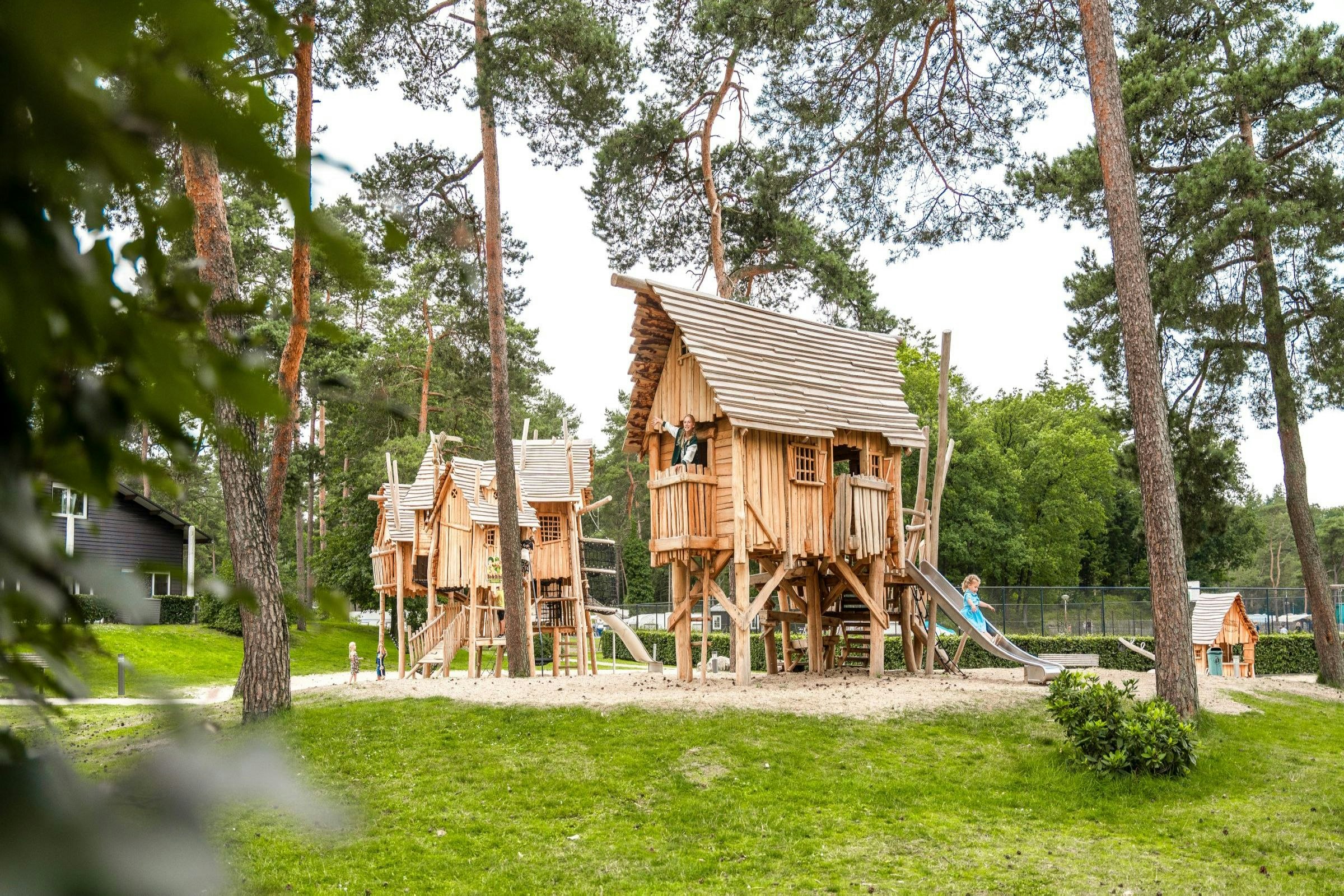 Holz-Spielhaus auf einem Campingplatz mit Spielgerüst, Rutsche und Holzstegen im Wald zwischen Bäumen.