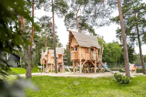 Holz-Spielhaus auf einem Campingplatz mit Spielgerüst, Rutsche und Holzstegen im Wald zwischen Bäumen.