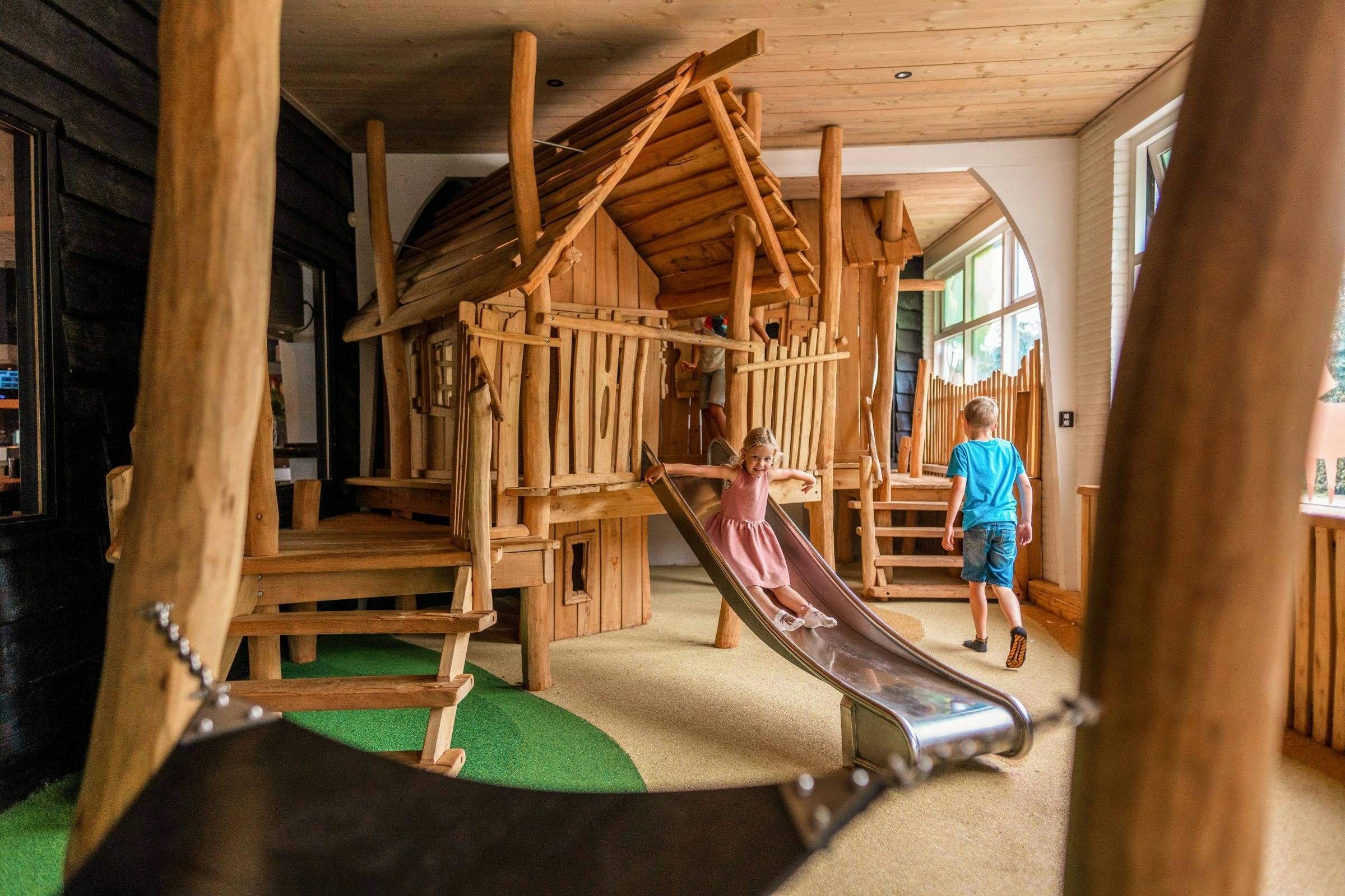 Kinder spielen im Holz-Spielhaus mit Rutsche, Geländer und Treppe, im Innenbereich mit Holzwänden und Fenstern.