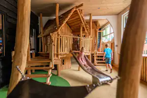 Kinder spielen im Holz-Spielhaus mit Rutsche, Geländer und Treppe, im Innenbereich mit Holzwänden und Fenstern.