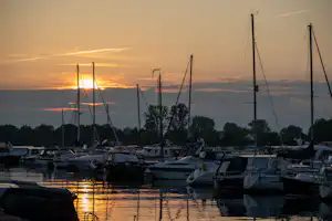 Hafen mit vielen Motorbooten und Segelbooten, hohe Masten und Taue im Vordergrund, Sonnenuntergang am Himmel.