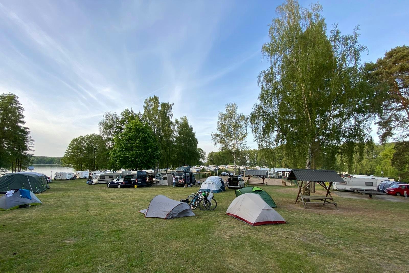 Zeltplatz mit mehreren Zelten und Wohnwagen auf einer Rasenfläche, Tische und Stühle am Rand, Bäume und Himmel im Hintergrund.